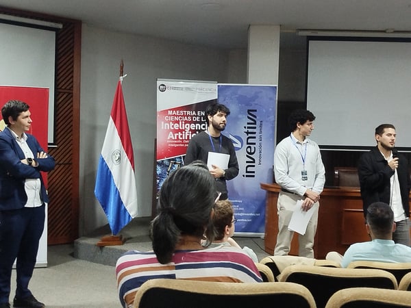 A group of men are standing on a stage during a presentation, with banners and the Paraguayan flag visible behind them and an audience in the foreground.