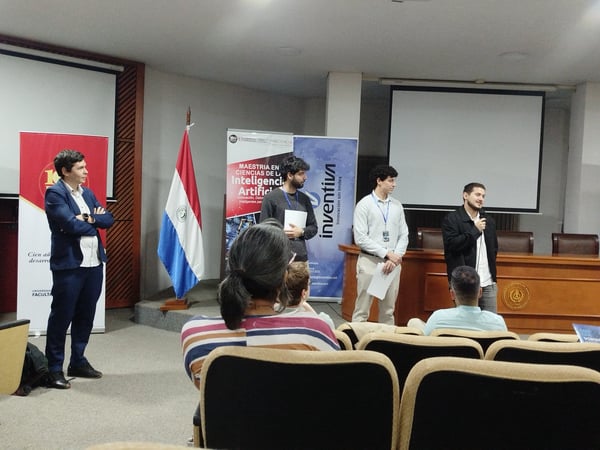 A group of people are gathered in an auditorium for a presentation about Artificial Intelligence, with several speakers standing at the front near banners and a Paraguayan flag.