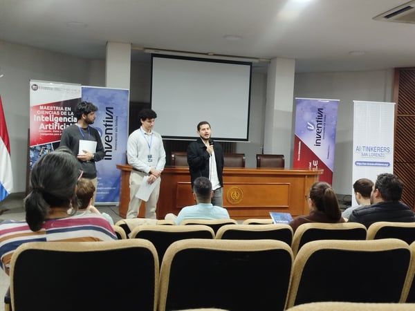 A man is giving a presentation with a microphone in front of an audience in an auditorium, flanked by two other men and several promotional banners.