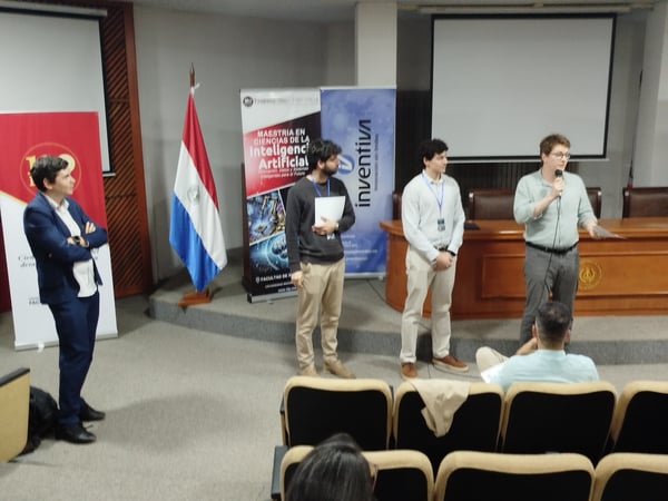 The image shows a group of men on a stage in an auditorium during a presentation, with one man speaking into a microphone. Banners for a Master's program in Artificial Intelligence and a Paraguayan flag are visible in the background.