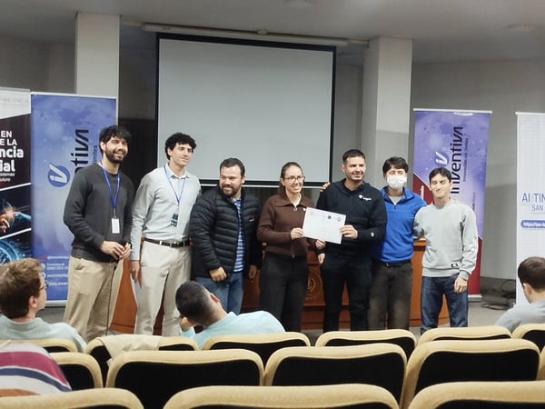 A group of seven people stands at the front of an auditorium, holding a certificate during an event with banners in the background.