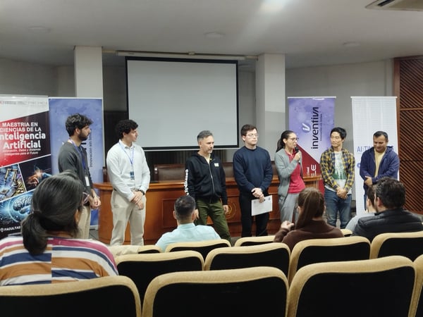 A group of people stands at the front of an auditorium during a presentation or event, with several banners visible and an audience seated in the foreground.