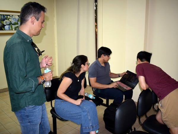 A group of four people are gathered in an indoor setting, focusing on a laptop screen held by one of them.