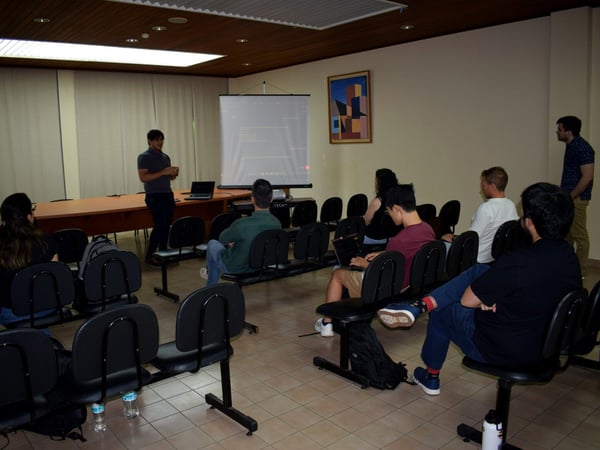 A man is giving a presentation to a small audience in a room with a projector screen and rows of chairs.