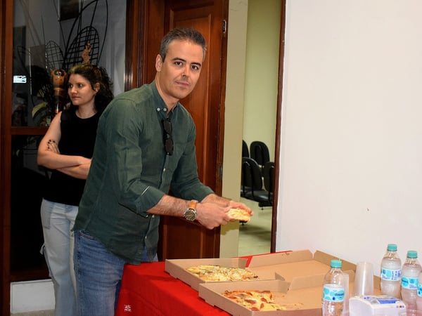 A man in a green shirt is taking a slice of pizza from a box on a table covered with a red cloth, while a woman stands behind him.