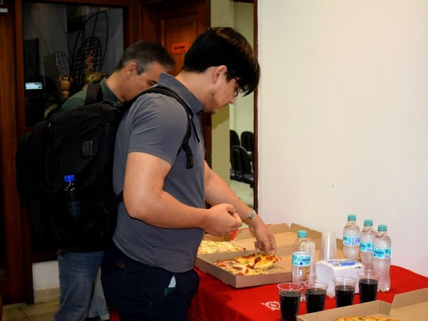 A man with a large black backpack reaches for a slice of pizza from a box on a table covered with a red cloth, which also holds water bottles and cups of soda.