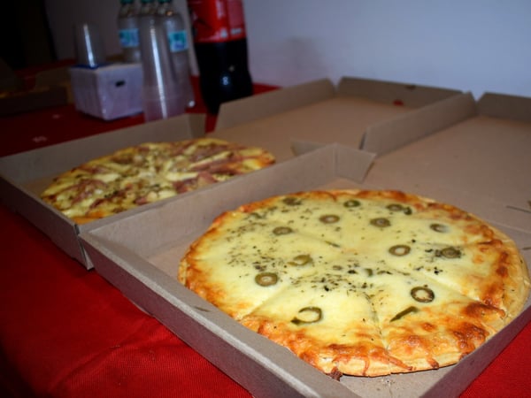 Two pizzas in open cardboard boxes are placed on a red tablecloth, with bottles of water and soda in the background.