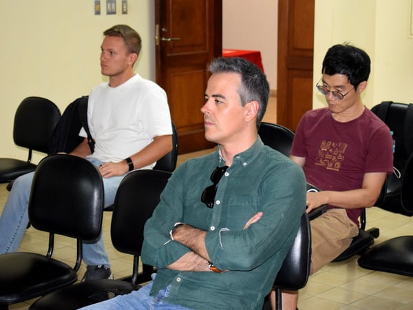 Three men are sitting in black chairs in what appears to be a classroom or meeting room, looking towards something off-camera.