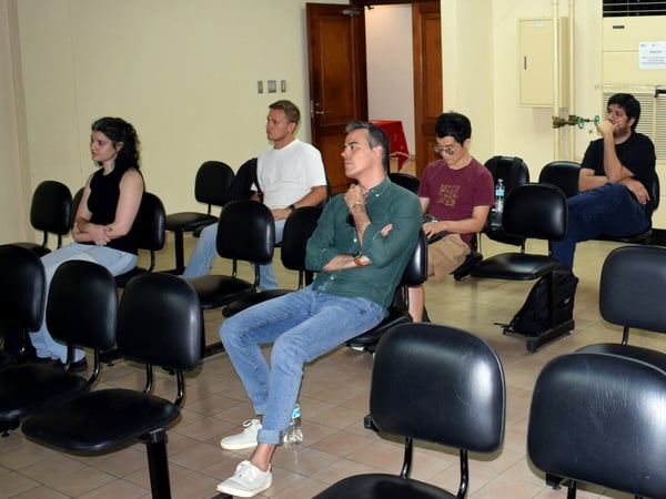 A group of five people are seated in rows of black chairs in a room, appearing to be attentively listening to a presentation or speaker off-camera.