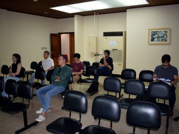 A group of people are seated in rows of black chairs within a room, appearing to attend a presentation or meeting.