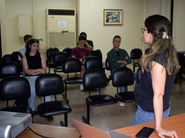 A woman stands in front of a small audience in a classroom or meeting room, likely delivering a presentation or leading a discussion.