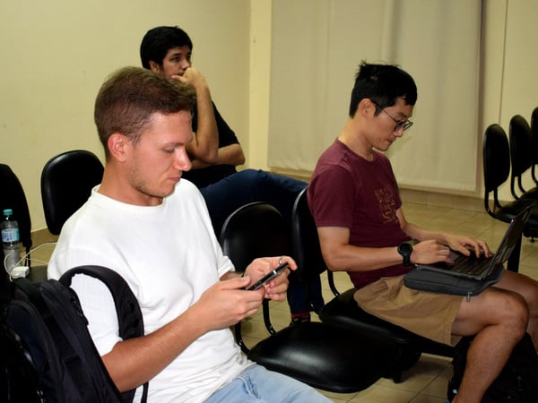 Three men are seated in an indoor setting; one is using a smartphone, another is typing on a laptop, and a third man is sitting behind them looking thoughtful.
