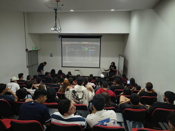 A photograph of a lecture hall where a presenter is showing a GitHub repository on a projector screen to a seated audience.