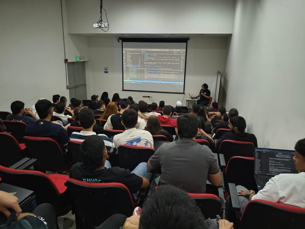A group of people are seated in a lecture hall, watching a presentation on a projector screen that displays computer code.