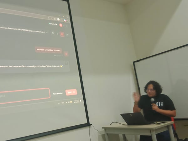 A person is giving a presentation in a room, holding a microphone and standing next to a laptop while a large screen displays a chat interface.