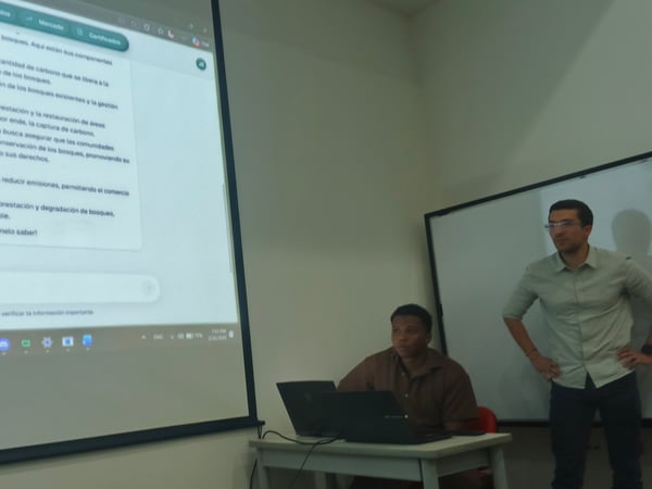 A man stands next to a large projection screen displaying a Spanish document about forest conservation and emission reduction, while another man sits at a desk with a laptop.