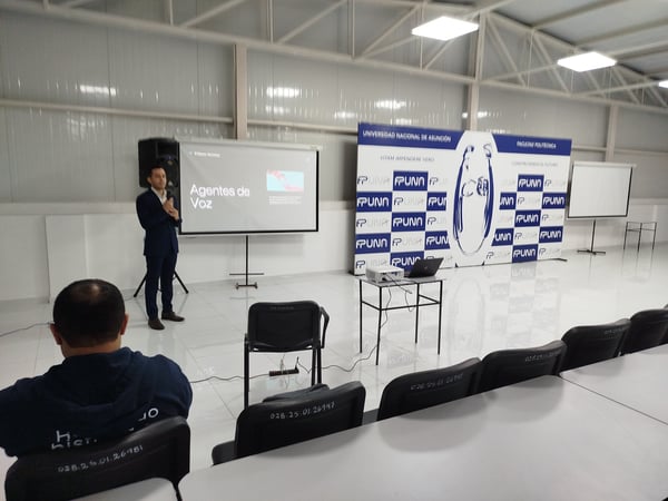 A man in a suit is delivering a presentation titled 'Agentes de Voz' in a spacious hall at the Universidad Nacional de Asunción, with a large institutional banner in the background.