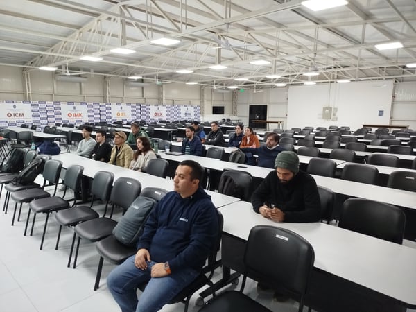 A group of people are seated at long white desks with black chairs inside a large, warehouse-like seminar hall with a metal truss ceiling.