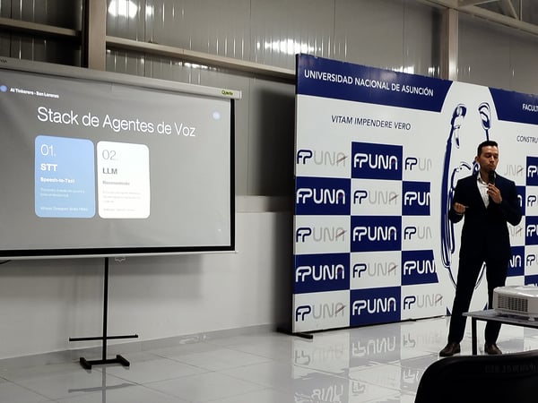 A man in a dark suit is delivering a presentation next to a projector screen displaying a slide about a 'Stack de Agentes de Voz' and a backdrop banner for the Universidad Nacional de Asunción.