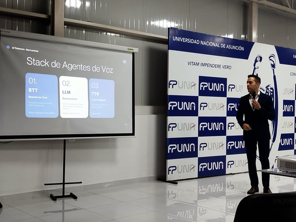 A man in a dark suit stands next to a projector screen presenting a slide about a 'Stack de Agentes de Voz' in front of a backdrop with the FPUNA logo.