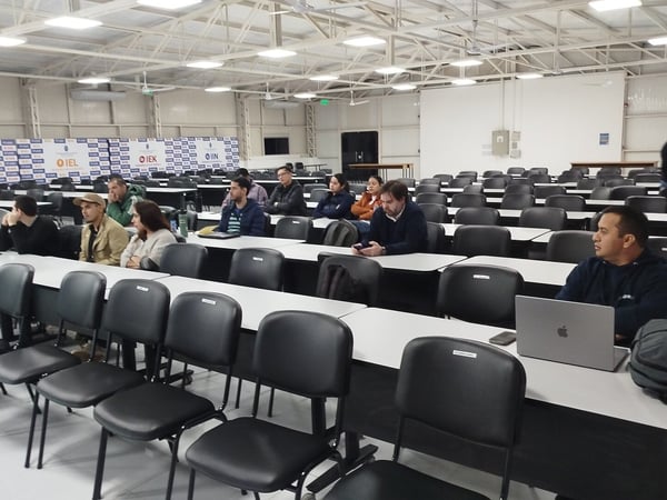An indoor photograph of a large lecture hall with rows of empty black chairs and white tables, where a small group of people are seated and waiting or listening.