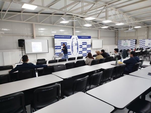An indoor photograph showing a presentation taking place in a large classroom or hall at the Universidad Nacional de Asunción. A presenter stands near a projection screen and university banners while a small audience sits at white tables listening.