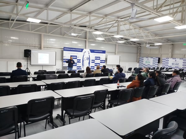 A speaker delivers a presentation to an audience seated at desks in a large university lecture hall.