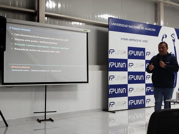 A man stands next to a projector screen presenting a slideshow in Spanish about vector spaces, with a University of Asunción backdrop behind him.