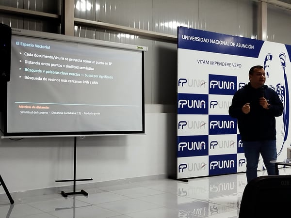 A man is presenting a slideshow about vector spaces next to a promotional banner for the Universidad Nacional de Asunción.