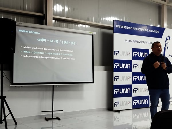 A man stands next to a projection screen displaying a slide on cosine similarity during a presentation at the Universidad Nacional de Asunción.