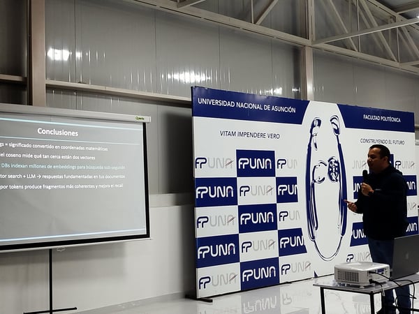 A photograph showing a man delivering a presentation in Spanish, standing next to a projector screen displaying conclusions and a large FPUNA banner backdrop.