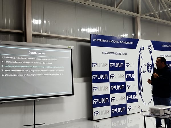 A man stands next to a projector screen displaying presentation conclusions in Spanish, with a large backdrop banner for the Universidad Nacional de Asunción in the background.