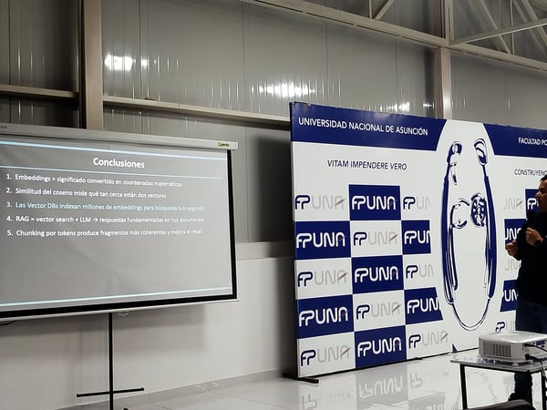 A photograph showing a presentation setup with a projector screen displaying conclusions about embeddings and RAG, next to a promotional backdrop banner for the Universidad Nacional de Asunción (FPUNA).