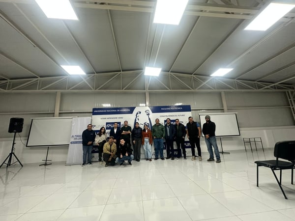 A group of thirteen people posing for a photo inside a spacious room with a high ceiling, standing in front of a large banner of the Universidad Nacional de Asunción.