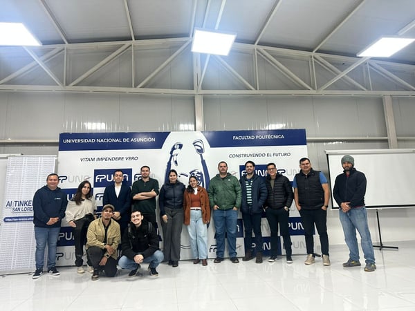 A group of thirteen people are posing for a group photo indoors in front of a blue and white banner for the Universidad Nacional de Asunción.