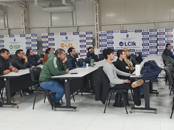 An indoor photograph showing a group of students or attendees sitting at desks in a classroom or lecture hall, attentively watching a presentation. The background features several banner backdrops representing different degree programs from FP-UNA, such as LCA, ISP, LEL, LCIk, and LCI.