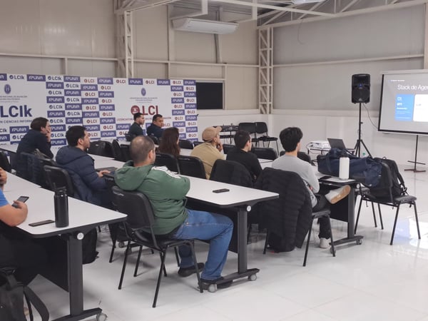 A photograph showing a presenter in a suit delivering a presentation titled 'Stack de Agentes de Voz' to an audience seated in a classroom or lecture hall.