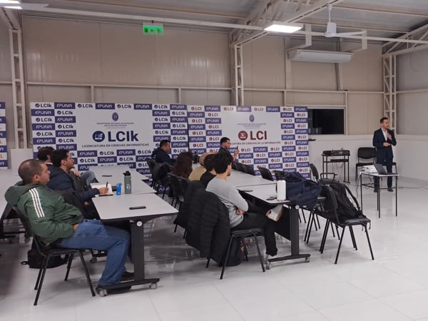 An indoor photograph showing a speaker giving a presentation on speech synthesis (TTS) to an audience seated at tables in a university hall.