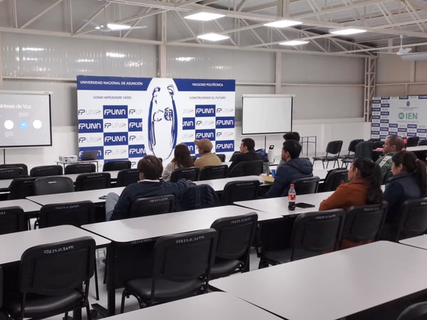 An indoor photograph showing a presentation in a classroom or lecture hall, with a speaker standing next to a projector screen and several audience members seated at desks.