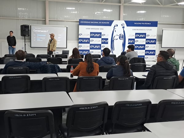 A presentation is taking place in a large hall, where a speaker stands at the front next to a projector screen while an audience sits at long tables facing them.