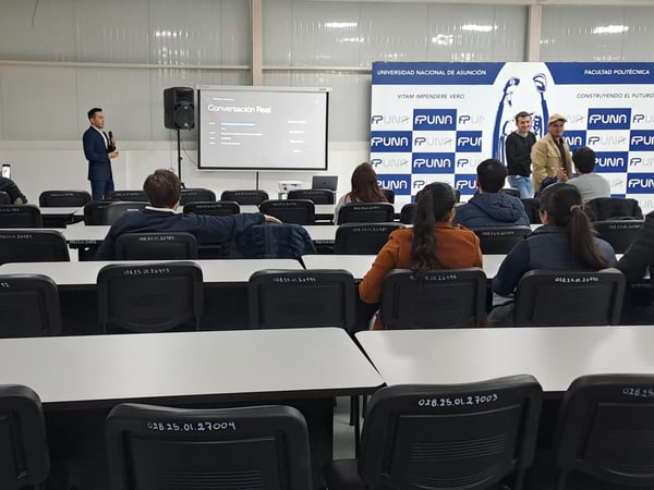 A photograph showing a presentation inside a university classroom or hall, with a speaker standing near a projector screen and an audience seated in rows facing forward.