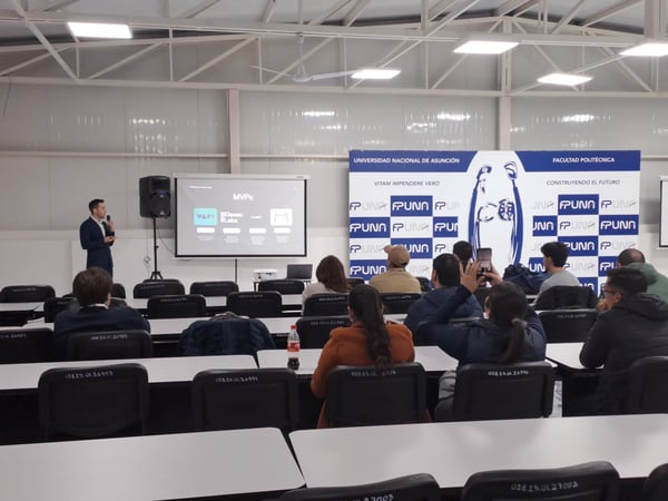 A presentation is taking place in a large university classroom or lecture hall, where a speaker stands at the front next to a projector screen showing 'MVPs' while the audience watches from their desks.
