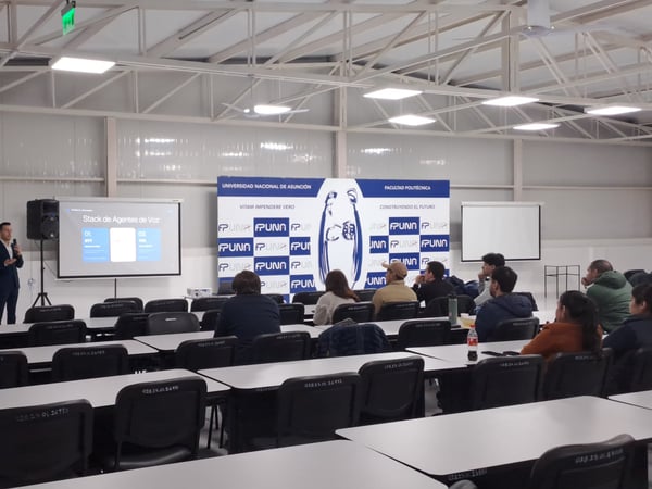 A photograph of a presentation taking place inside a large classroom or hall, where a speaker stands at the front next to a projector screen displaying 'Stack de Agentes de Voz' to an audience.