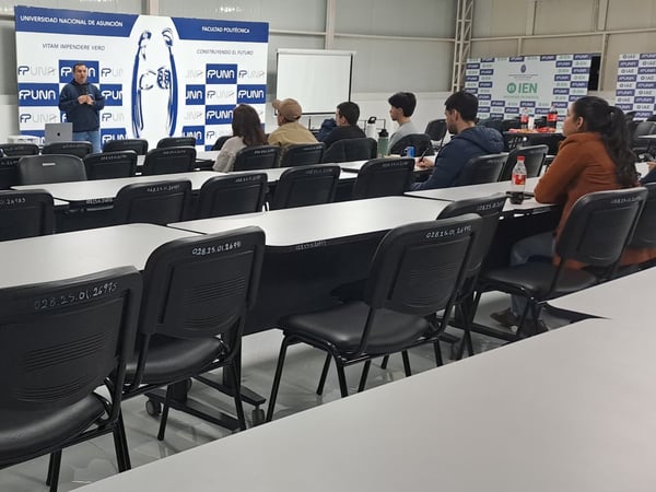 An indoor photograph showing a speaker presenting to a small audience seated at desks in a classroom or lecture hall at the Universidad Nacional de Asunción.