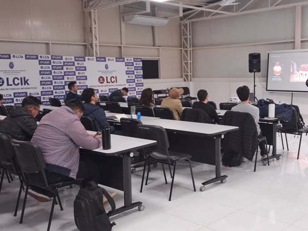 A man is giving a presentation to a seated audience in a large classroom or hall at the Universidad Nacional de Asunción, with banners displaying university logos in the background.