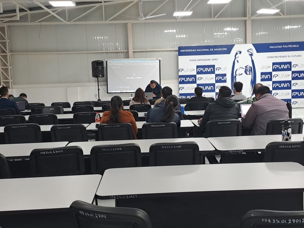 An indoor photograph of a university lecture hall where students are seated at desks facing a presenter and promotional banners.