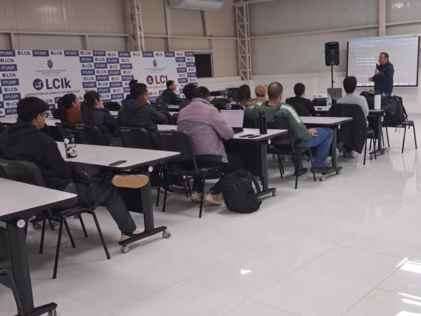 An indoor photo of an academic presentation, showing an audience seated at desks watching a speaker stand near a projection screen.