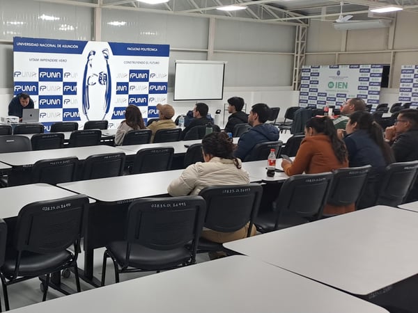 An indoor shot of a university classroom or presentation hall where an audience is seated at rows of desks, watching a presenter set up a slideshow on a projector screen.
