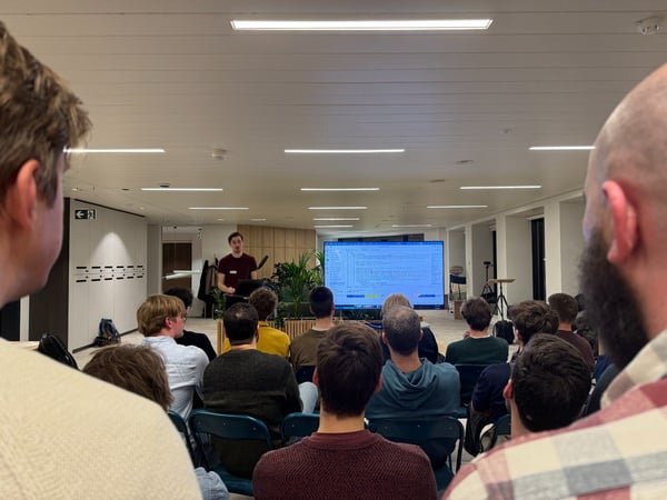 A group of people are seated in an indoor space, attending a presentation where a speaker is standing next to a large screen displaying code.