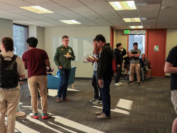 A group of people are socializing and eating in a modern office common area with a whiteboard and large windows.